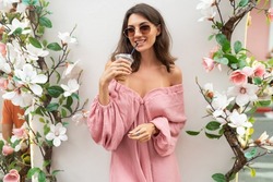 Young beautiful smiling cute romantic woman in trendy summer pink dress. Carefree woman posing in the street near white wall with flowers. With cup of iced coffee latte
