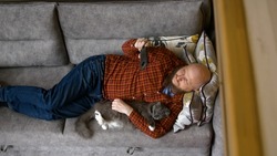 Shooting from above, man lies on couch half-side with cat under his arm, scratches his pet and watches movie on phone. Caucasian man in plaid red shirt is resting on sofa with a cat and a smartphone.