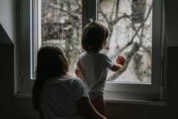 Rainy day. Little toddler boy with his sister looking outside through  wet window from his room missing mother.  Siblings love, care and support. Lifestyle. Quarantine and isolation period at home