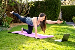 Happy smiling woman practicing pilates lesson online in garden outdoors during quarantine. Doing sport at home following guide or online tutorial or trainer instructions on laptop. 