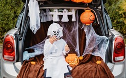 Trick or trunk. Trunk or treat. Happy child in ghost costume having fun celebrating Halloween party in decorated trunk of car. New trend and alternative safe outdoor celebration of traditional holiday