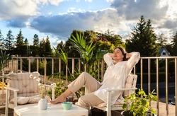 Beautiful young girl relaxing and enjoying sun while having tea, sitting at balcony at sunlight at summer. Slow living, tranquil moment, mental health. Backyard terrace vacation.
