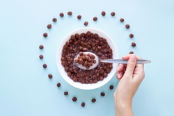 Hand with a spoon over a bowl with chocolate cereal balls on blue background.Dry breakfast cereal top view. Heart shape from flakes. Concept for a healthy diet.