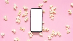Flat lay scattered popcorn with mobile phone with blank screen on pink background. Mockup phone with white copy space. Smartphone application for online cinema, films and media. 