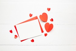 Greeting card mockup, white blank card with paper envelope and red parer hearts on wooden table background. Valentine's day greeting card, flat lay, top view