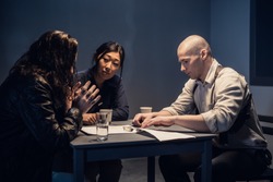 A police detective at the station and his partner, an asian woman, present evidence to a male suspect in an interview room