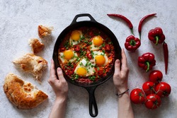 Shakshuka with pita. Hands holding a frying pan. Traditional dish of the Middle East fried eggs with tomatoes, sweet peppers, chili, vegetables and herbs on a white background. View from above. 
