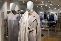 Two women's mannequins in the window of a large outerwear store. Autumn winter coat made of wool of natural color with a belt and winter long knitted dress. Shop hall in the background