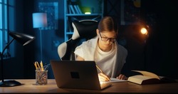 Young caucasian teenage girl using laptop, studying doing school lessons late at night. College student browsing internet, searching information, writing notes.