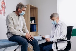 Elderly patient wearing medical mask talking to male family therapist during regular doctors appointment. Senior man, suffering knee pain, taking care of his health during coronavirus pandemic.