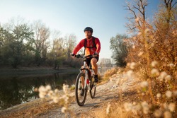 Happy young bearded man wearing red long sleeve cycling jersey and a backpack enjoying late afternoon mountain bike ride through the forest by river on a clear autumn day