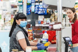 A young woman in a medical mask and gloves, working at the checkout in a supermarket. In the background, the buyer is blurred. Concept of coronovirus, protection from infection and industrial crisis