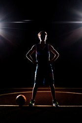 Basketball. The silhouette of a teenage Boy in blue sportswear confidently poses with his hands on his hips. The ball is on the ground. Black background with spotlights. Concept of sports games