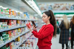 A young woman scans the QR code on a package of yogurt. In the background, a supermarket with visitors in a blur. The concept of modern technologies.