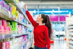 A young Caucasian woman reaches to the top shelf for a pack of sanitary pads. Side view. Corridor of shop on the background. The concept of purchasing hygiene products and cosmetics.
