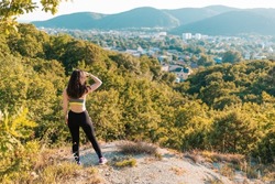 Young athletic woman looking into the distance, standing on a rocky ledge. In the background there is a forest and a view of the city. Sports, fitness and healthy lifestyle concept. Back view.