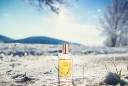 Rectangular transparent glass bottle of golden perfume on ice. A snow-covered blurred landscape in the background. Perfume and Fragrance template concept.