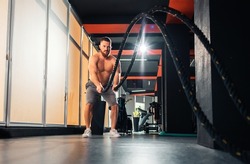 Athletic caucasian young man with battle rope doing exercise in fitness gym. Keeping fit with cross training.
