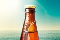 Close-up of the neck of wet glass bottle of dark beer against the background of the ocean and the sky. Inside it drowning hand. Alcoholic beverages on vacation.