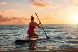 Plus sized beautiful Caucasian woman in red swimsuit swimming sitting on a sup board. Sunset on the background. Back view. The concept of sports and summer vacation.