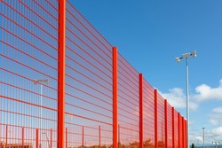 Modern fencing of the sports ground with a metal red fence. Lighting devices in the background