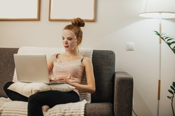 Caucasian woman with red hair is wearing a cleaning mask on her face while sitting on the sofa and using a laptop