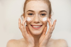 Beautiful ginger girl applying special cream on her freckled face while posing on a beige wall