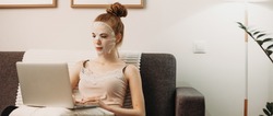 Red haired caucasian lady sitting on the sofa and working at the computer while wearing a paper special mask for face skin