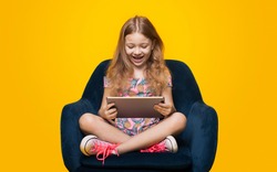 Lovely blonde girl playing with a tablet on the armchair posing with a yellow studio wall on background