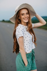 Caucasian redheaded woman wearing a hat posing on a road over picturesque landscape. Beautiful girl. Pretty young woman.