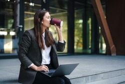 Young woman working online on laptop computer sitting outside the office building wearing wireless headphones enjoying a takeaway coffee. Freelancer working