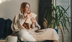 Thoughtful and cozy red-haired woman sitting with her legs crossed and taking notes on notebook. Relaxation lifestyle concept