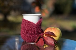 A white cup of coffee is wrapped in a burgundy scarf. The blue sky is reflected in the glass surface of the table. Red leaf and yellow apple. Natural background of autumn garden trees. bokeh.