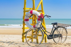 Old retro black bicycle with flowers bouquet beside Life ring for life safety on yellow lifeguard stand station or lifeguard chair protecting the safety of tourist on the empty beach in sunshine.