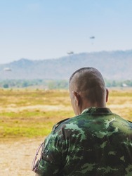 Back view of Asian army soldier stands to explore the landscape in field. Command class soldier stands to watch and inspect the conditions of the maneuver in open terrain with parachute in background.
