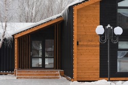 The house is in a modern style. Eco-friendly housing. Combination of wood and metal. Entrance door, panoramic windows. It's winter, there's snow and trees all around.