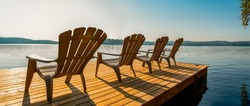 Row of Adirondack chairs -patio - deck chairs on wooden dock with sunset or sunrise -cottage life. Banner - panoramic