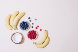 Bananas, coconut, raspberries and blueberries on neutral beige background. Harsh shadows. Top view, flat lay, copy space