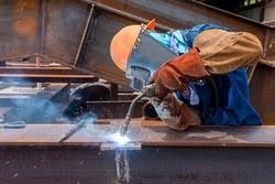 Welder is welding add joint h-beam for steel structure work with process Flux Cored Arc Welding(FCAW) and dressed properly with personal protective equipment(PPE) for safety, at industrial factory.