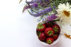 Fresh strawberries in a bowl with bouquet of summer flowers on white table. Top view, copy space. Midsummer still life