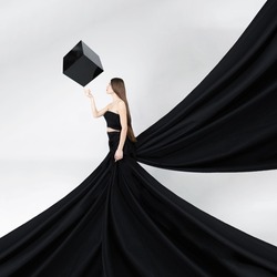 Fashion, fine art, fantasy concept. Beautiful long brown hair woman with huge black dress and flying trail like wings touching big black levitating cube in white background. Image with motion blur