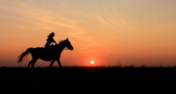 Horseback woman riding on galloping horse with red rising sun on horizon. Beautiful colorful sunset header background with equine and girls silhouette. 