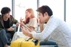 Asian young man holding and looking at mobile phone while waiting airline flight to travel, showing uncomfortable and worried expression, having of signs of text neck syndrome.