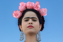 Portrait of look alike popular Mexican artist Frida Kahlo with her fashion statement with bright red floral head piece,  ornament and popular Unibrow over a blue background.