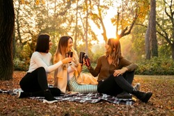 Group of three female friends smiling, sitting on picnic blanket in the park and drinking coffee, having a good time, fall season 