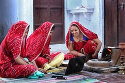 Three traditional Indian young married women working in traditional kitchen on laptop. Using technology in rural households. Women cook food and learn technology. Rural women using Laptop. 