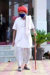 An old wrinkled indian rural man in red turban walking wearing mask. Protection from covid-19 pandemic. Indian rural traditional male dress now includes mask. Mask a latest fashion statement for old.