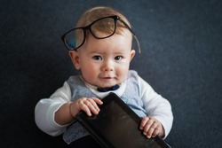intelligent Cute baby genius who looks like a little student well dressed in vest and bow tie with glasses holding tablet for working and education on grey background