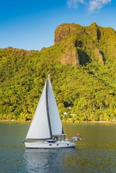Beautiful blue water luxury sailing yacht leaving Cook's Bay, Moorea, French Polynesia, with full sails up and the majestic volcanic mountains in the background, green tropical vegetation and blue sky