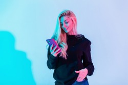 Happy beautiful girl with a smile in a fashion black hoodie and blue jeans holds a smartphone and looks at the application in the studio on a colorful neon pink lights. Creative modern female portrait